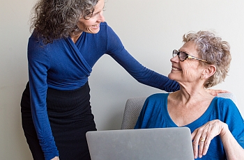 A smiling woman standing next to an smiling old woman who is using her computer